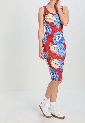 Frau trägt ein rotes ärmelloses Kleid mit großen blauen und weißen Blumenmustern, kombiniert mit weißen Sneakers und Socken.