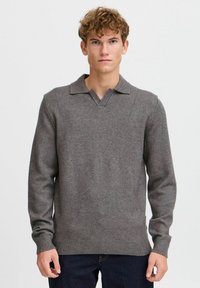 Grauer Strickpullover mit V-Ausschnitt und Kragen. Lange Ärmel und gerippter Saum. Weicher Stoff, schlichtes Design. Über dunklen Jeans getragen.