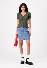 Maglietta verde oliva a coste con maniche corte a palloncino, abbinata a una gonna di jeans azzurra chiara. Gli accessori comprendono una borsa rossa e scarpe con plateau nere.