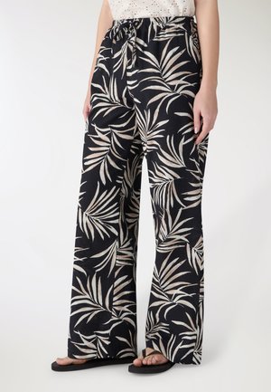 Donna che indossa pantaloni neri a gamba larga con stampa di foglie beige, vita elastica e infradito nere, in piedi su uno sfondo bianco semplice.