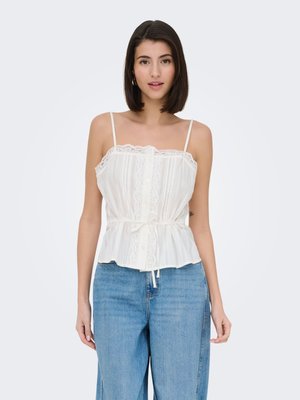 Woman with straight dark hair wearing a white lace-trimmed sleeveless top and high-waisted blue jeans against a plain background.