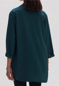 Długi, ciemnoniebieski tunikowy shirt wykonany z teksturowanej tkaniny w kratkę. Posiada podwinięte rękawy oraz klasyczny krój kołnierzyka.