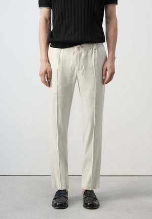 Homme portant un pantalon droit couleur crème avec pli central, un haut noir en maille à manches courtes et des mocassins en cuir noirs, debout sur un sol gris.