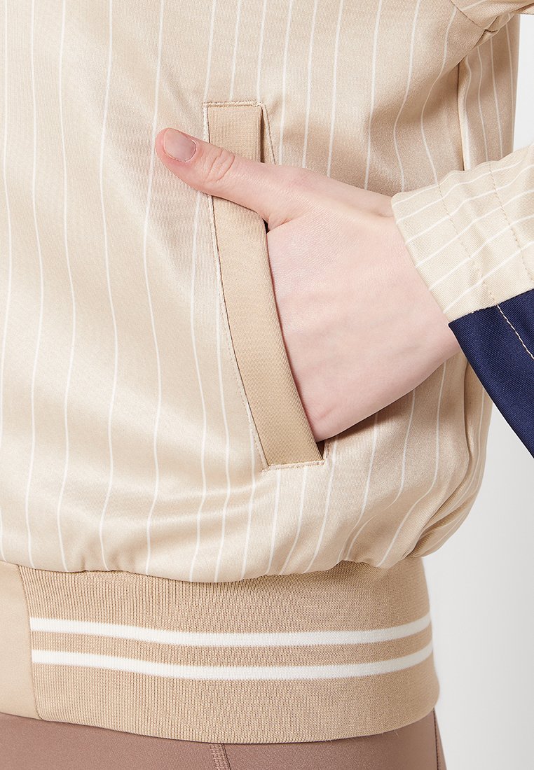 Chaqueta bomber de satén beige con rayas verticales, que presenta un bolsillo lateral y un dobladillo acanalado con rayas blancas, mostrando una textura suave.