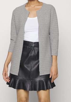 Femme portant un haut blanc sans manches, un cardigan gris clair côtelé ouvert devant, et une jupe en cuir noire avec un ourlet à volants.