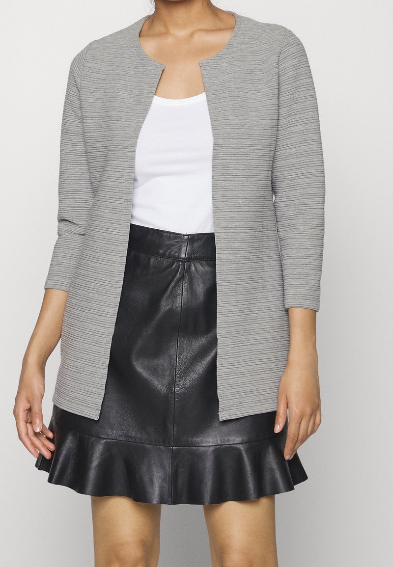 Femme portant un haut blanc sans manches, un cardigan gris clair côtelé ouvert devant, et une jupe en cuir noire avec un ourlet à volants.