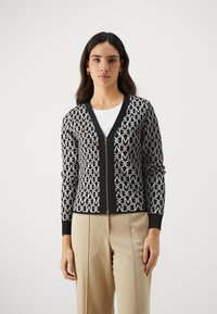 Svart og hvit glidelås-foret cardigan med et gjentakende logo-mønster, V-hals og ribbede mansjetter. Sammen med lyse bukser.