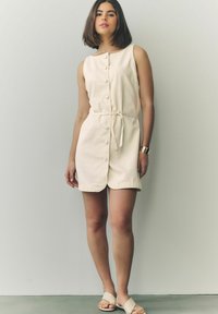 Robe en mélange de coton beige avec un devant boutonné, design sans manches, ceinture ajustée à la taille et ourlet arrondi. Portée avec des sandales crème.