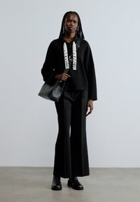 Kvinde iført sorte bukser med vide ben, sort sweater med hvide mønstrede snore, sorte støvler og bærende en sort håndtaske.