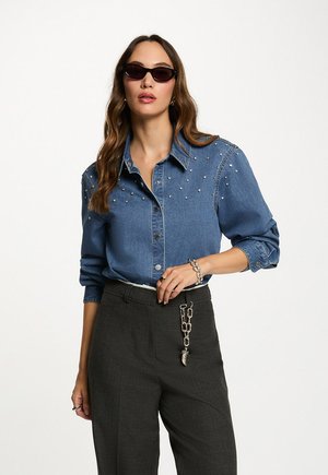 Camisa de denim con acentos de perlas en los hombros, cuello clásico y cierre de botones, combinada con pantalones de sastre grises de cintura alta y gafas de sol.