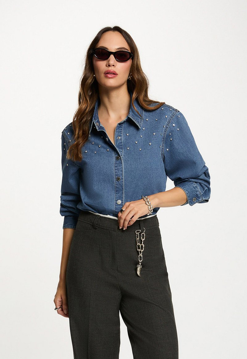 Camisa de denim con acentos de perlas en los hombros, cuello clásico y cierre de botones, combinada con pantalones de sastre grises de cintura alta y gafas de sol.