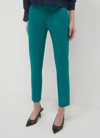 Pantalon fuselé couleur sarcelle en tissu lisse, avec une taille mi-haute et une silhouette élégante. Porté avec des escarpins noirs à bout pointu.