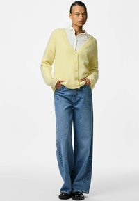 Gul fluffig cardigan med v-ringning, med texturerad stickning och fem knappar. I kombination med vida blå jeans och svarta skor.