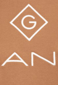 Hnědá tkanina s hladkým povrchem, na které se nacházejí velká bílá vyšitá písmena "G" a "AN" uvnitř diamantového tvaru.