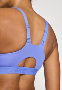 Paarse sportbeha gemaakt van gladde stof, met een rugontwerp met uitsnijding en verstelbare bandjes. Branding zichtbaar op de band.