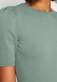 T-shirt med kort ärm och rund hals i mjukt mintgrönt tyg. Har samlad detalj vid axlarna och en slät, figurnära textur.