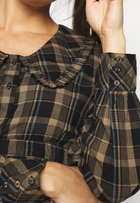 Blouse à carreaux marron et noir avec un col à volants et des manches bouffantes. Présente des boutons noirs sur le devant et aux poignets.