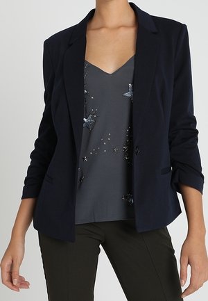 Vrouw draagt een marineblauwe blazer over een donkergrijze mouwloze top met ster- en maanversieringen, gecombineerd met een donkere broek.