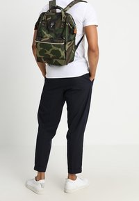 Henkilö, jolla on päällään valkoinen paita, mustat housut, valkoiset lenkkarit ja vihreä kamouflage-reppu, seisoo kädet taskuissa.