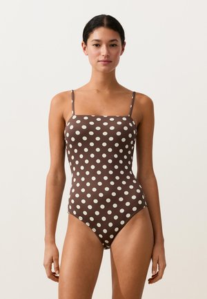 Žena stojaca čelom v hnedých jednodielnych plavkách s bielymi bodkami a tenkými ramienkami na jednotnom pozadí.