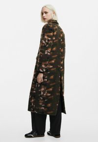 Lang, grøn camouflagefrakke med nuancer af brune og beige, lavet af tekstureret stof; har en klapkrave og sideslidser.