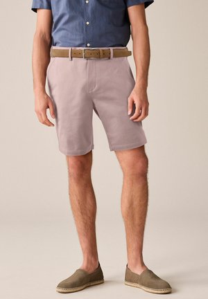 Pantaloni scurți roz deschis, cu o textură netedă, având o curea maro și un tricou albastru cu mânecă scurtă. Poartă espadrile verzi-măslinii.