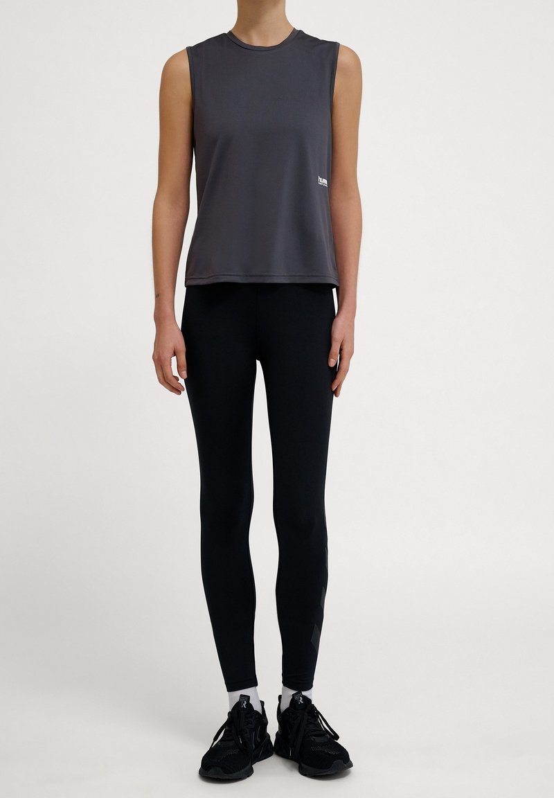 Haut de sport gris sans manches avec un col rond, associé à des leggings noirs et des baskets noires. Design minimaliste sans motifs notables.