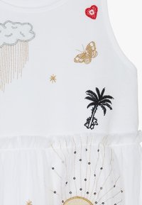 Biała sukienka zdobiona różnorodnymi kolorowymi haftami: srebrna chmurka, złoty motyl, czarna palma, czerwone serce oraz słońce z koralikowymi akcentami.