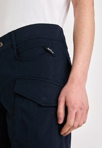 Pantalons cargo marine avec de multiples poches, détails cousus et fermeture à bouton brillant. Le tissu a une texture lisse.