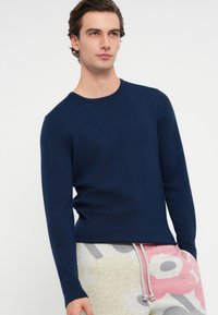 Mladý muž v priliehavých námořnicky modrých dlhých rukávoch a svetlých teplákoch s veľkým ružovým a šedým kvetinovým vzorom.