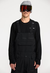 Zwarte sneeuwoverall met verstelbare schouderbanden, ritsvak, gecombineerd met een zwart lange mouwen shirt en gebreide muts, met reflecterende goggles.