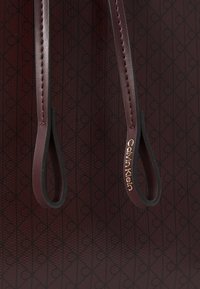 Calvin Klein torba u tamnocrvenoj koži s reljefnim uzorkom srca. Ima glatke ručke s zlatnim logotipom. Teksture površine.