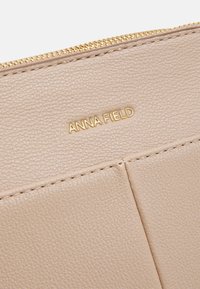 Beige pung i syntetisk læder med guld "ANNA FIELD" logo, med en tekstureret overflade og lynlås. Dekoreret med syninger.