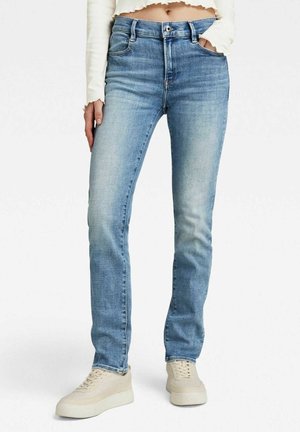 Light blue slim-fit jeans worn with beige sneakers and a cropped long-sleeve white top, one hand in pocket.