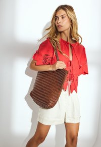 Braune gewebte Tragetasche mit einer weichen, flexiblen Textur, kombiniert mit einer leuchtend roten Bluse mit aufwendigen Ausschnitten und hellen Shorts.