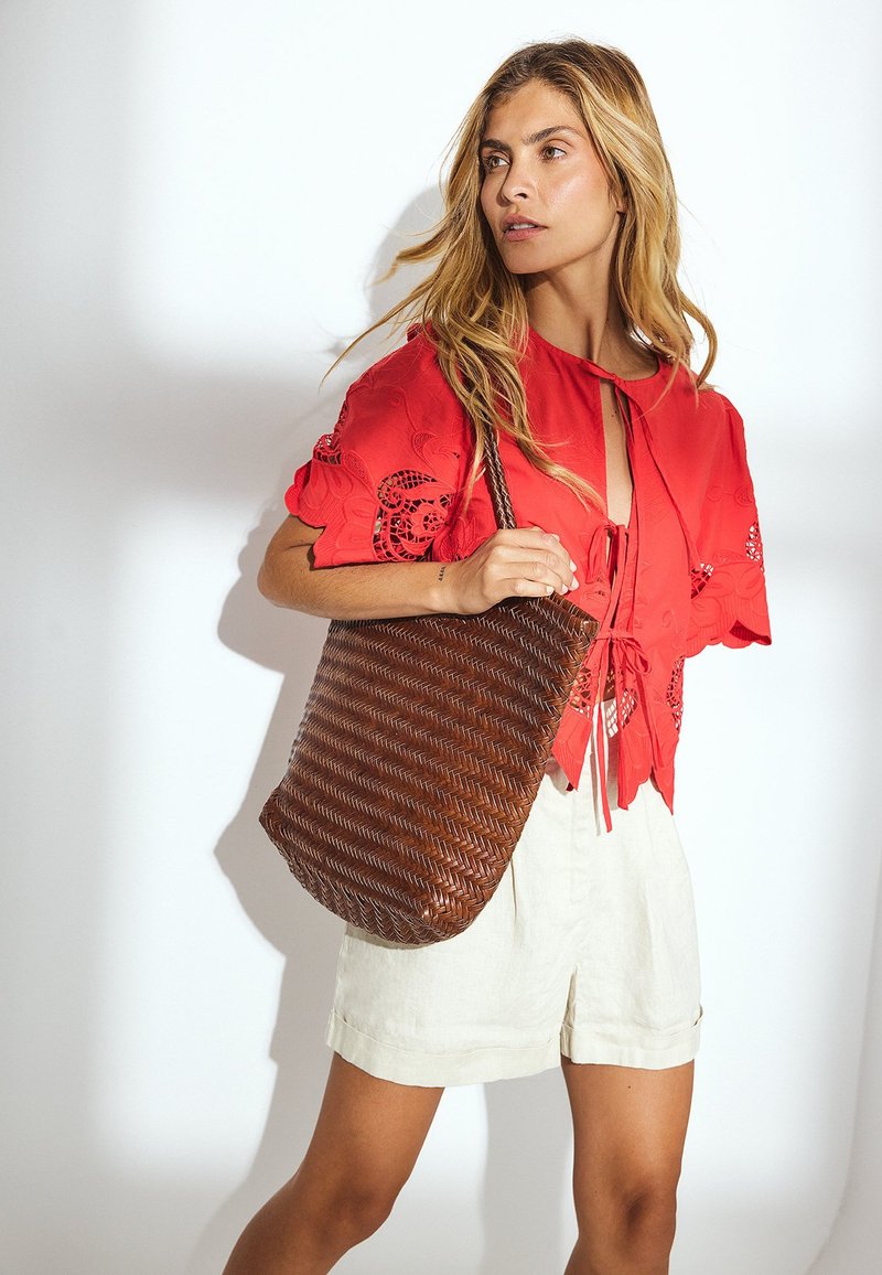 Braune gewebte Tragetasche mit einer weichen, flexiblen Textur, kombiniert mit einer leuchtend roten Bluse mit aufwendigen Ausschnitten und hellen Shorts.