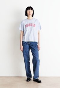 Hellblaues Baumwoll-T-Shirt mit "CALVIN KLEIN" in rotem Text, kombiniert mit dunkelblauen Jeans, die einen lockeren Schnitt und ein minimales Design aufweisen.