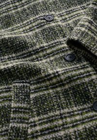 Wollmantel mit grünem, schwarzem und weißem Hahnentritt-Muster, ausgestattet mit schwarzen Knöpfen und einem Reverskragen. Strukturiertes Gewebe mit sichtbaren Nähten.