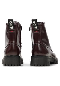 Par de botas de cuero marrón oscuro a la altura del tobillo con suelas de goma negra gruesa, con tiradores de cremallera traseros etiquetados como "HUGO," vistas desde la parte trasera.