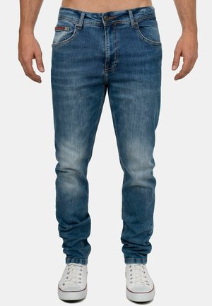 Jeans in denim blu con un design aderente, caratterizzati da tasche frontali, sfumature sottili e dettagli a doppia cucitura. Abbinati a sneakers bianche.