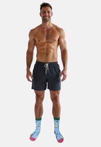 Bañador de hombre en color azul marino con cintura ajustable, combinado con calcetines coloridos que presentan patrones de frutas sobre un fondo azul claro.