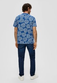 Niebieska koszula krótki rękaw z białym wzorem liści, strukturowana tkanina, okrągły dekolt. Połączona z ciemnoniebieskimi dżinsami i białymi trampkami.