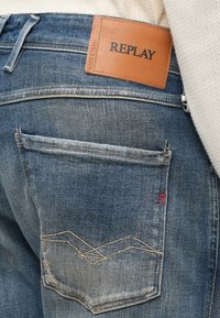 Μπλε τζιν παντελόνια από denim με δερμάτινο ετικετάκι με την ένδειξη "REPLAY." Διακρίνονται εμφανή σχέδια ραφών και ένα μικρό κόκκινο "R" στην πίσω τσέπη.