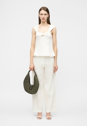 Vrouw draagt een wit mouwloos topje en broek met wijde pijpen, houdt een grote donkergroene gevlochten handtas in haar rechterhand.