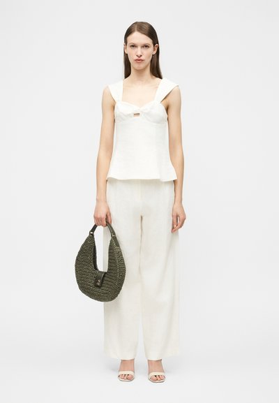 Woman wearing white sleeveless top and wide-leg pants, holding a large dark green woven handbag in her right hand.