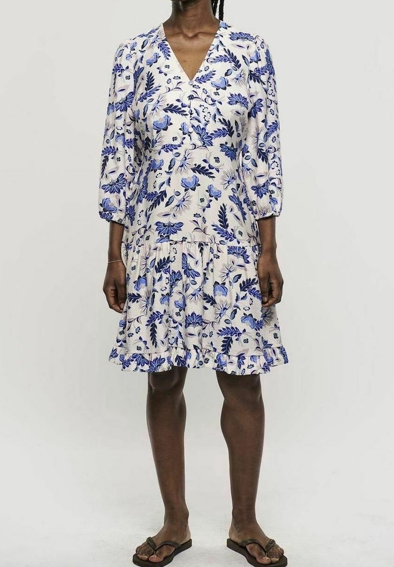Femme portant une robe blanche mi-longue avec des motifs floraux bleus, des manches trois-quarts bouffantes et des sandales tongs, debout devant un fond uni.