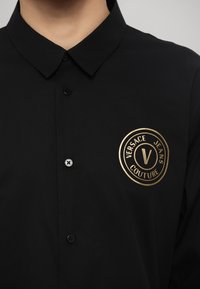 Musta nööpidega särk, millel on kuldne ringikujuline logo ja tekst "VERSACE JEANS COUTURE". Sile kangas klassikalise krae ja mustade nuppudega.