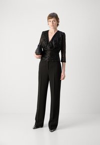 Schwarzes Paillettenoberteil mit tiefem V-Ausschnitt und Dreiviertelarm, kombiniert mit hochgeschnittenen schwarzen Hosen und spitzen Schuhen.