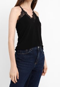 Svart ärmlös linne med spetsdetaljer, V-ringning och justerbara band, samsatt med högmidjade blå denimjeans.