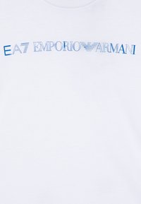 Bela bomba majica iz bombaža z modrim grafičnim napisom "EA7 EMPORIO ARMANI" in stiliziranim logotipom, gladka tekstura in krožni izrez.